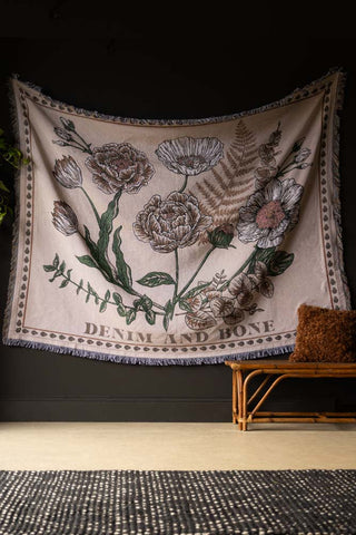 The Denim & Bone Scandi White Woven Cotton Throw displayed on a black wall above a wicker bench, cushion and a plant.