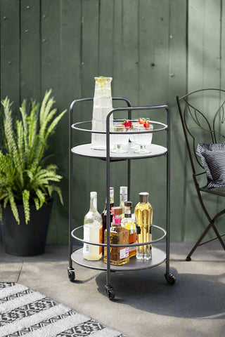 A grey outdoor drinks bar cart in a courtyard. The bar cart is filled with wine glasses and alcohol bottles.