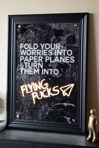 The Flying Fuck Framed Neon Wall Light with a black frame styled on a black sideboard.