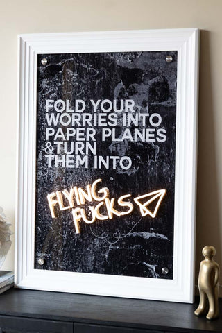 The Flying Fuck Framed Neon Wall Light with a white frame styled on a black sideboard.
