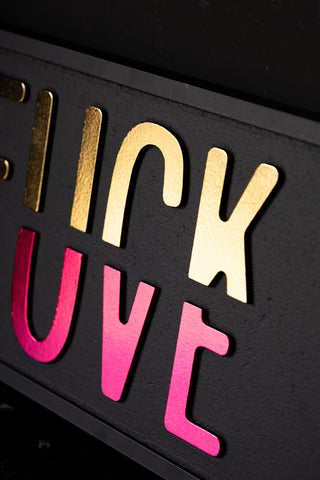 Close-up of the Fuck vs Love You Wall Sign leaning against a black wall.