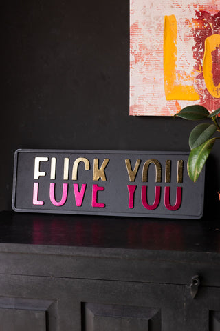 The Fuck vs Love You Wall Sign displayed leaning against a black wall on a sideboard, style with another piece of artwork and a plant.