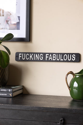 The Fucking Fabulous Sign displayed on a neutral wall with a framed artwork, above a black sideboard with a jug, potted plant and some books.
