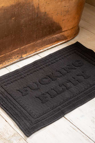 Black bath mat with the phrase 'fucking filthy' displayed on a wooden floor in front of a metal bath.