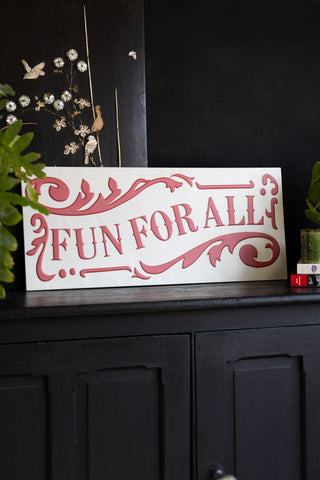 Decorative sign with 'Fun for All' text on a dark surface with plants.