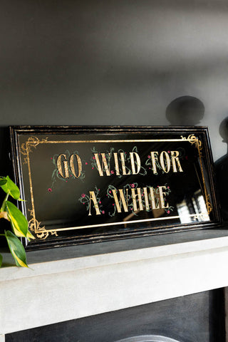 A styled image of the Go Wild For A While Decorative Mirror on a black fire place mantelpiece.