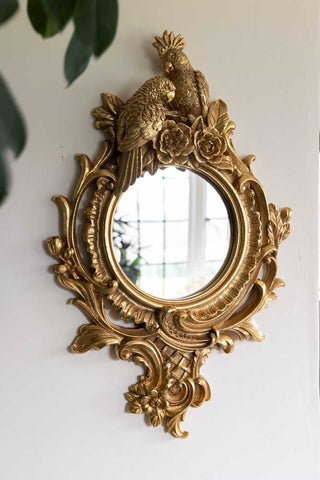The Gold Parrot Mirror displayed on a white wall with leaves hanging in the shot.