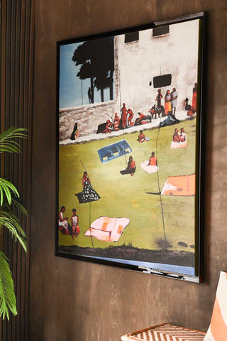 Lifestyle image of the HKliving Endless Summer Framed Artwork displayed on a dark wood wall, styled with a plant, rattan bench and cushion. 