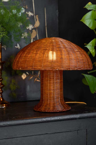Lifestyle image of the HKliving Rattan Toadstool Table Lamp styled on a black sideboard with greenery and home accessories.