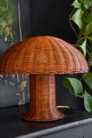 Lifestyle image of the HKliving Rattan Toadstool Table Lamp styled on a black sideboard with plants in the background. 