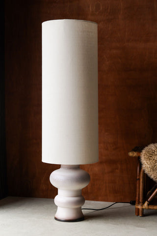 Lifestyle image of the HKliving White/Brown Retro Reactive Glaze Lamp displayed in front of a dark wooden wall next to a wicker bench with fluffy rug on. 