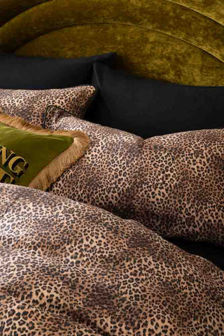 Detail image of the Leopard Love Duvet Cover and Pillow Case Set styled on a bed with a velvet headboard, with black pillows and a fringed cushion.