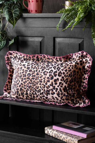 The Leopard Print Cushion With Pink Scalloped Edge styled on a black bench in front of a sideboard, with some books, plants, a mug and candle.