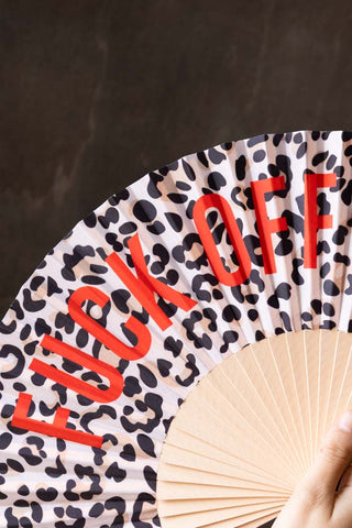 Close-up of the Leopard Print 'Fuck Off' Wooden Hand Held Fan in front of a dark wooden wall being held by a model's hand.