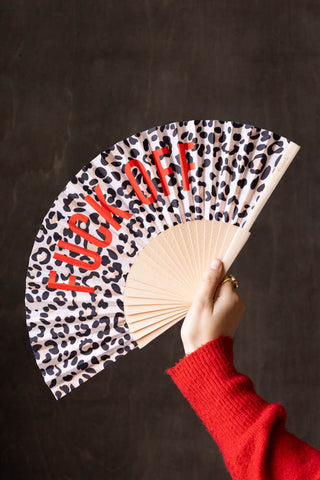 The Leopard Print 'Fuck Off' Wooden Hand Held Fan being held by a model in front of a dark brown wall.