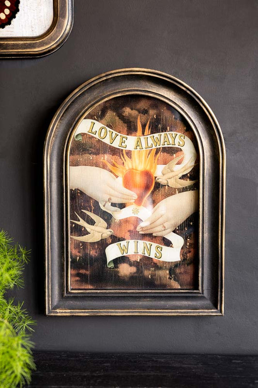 The Vintage-Style Love Wins Framed Wall Art displayed on a dark wall next to another artwork and a plant.