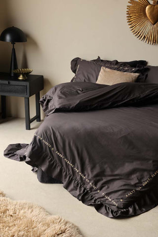 Lifestyle image of the Charcoal Grey Mega Frill Duvet Cover and Pillowcase Set styled on a bed in a neutral bedroom with a bedside table, lamp, rug and various other home accessories. 
