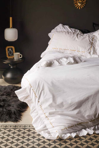 Lifestyle image of the White Mega Frill Duvet Cover and Pillowcase Set displayed on a bed in front of a black wall, styled with a light, side table, rug and other decorative accessories. 