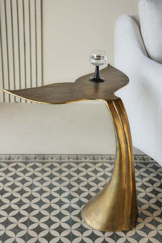 Lifestyle image of the Mermaid Tail Side Table with a black wine glass on the top, displayed on a geometric floor next to a sofa. 