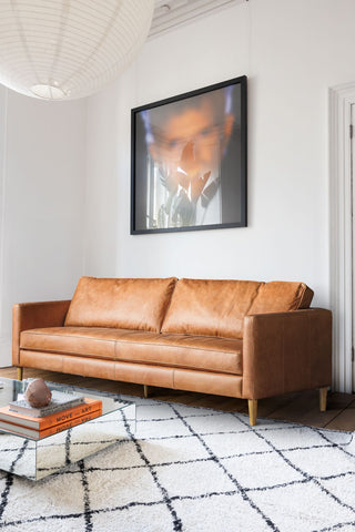 The Mid-Brown Vintage-Style Leather 3-Seater Sofa styled in the corner of a white living room, with a large print, coffee table, rug, pendant light and some accessories.