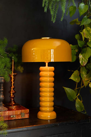 Lifestyle image of the Modern Metal Mustard Table Lamp displayed on a black sideboard surrounded by various other home accessories and plants.