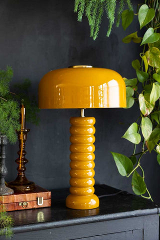 Lifestyle image of the Modern Metal Mustard Table Lamp displayed on a black sideboard surrounded by various other home accessories and plants.