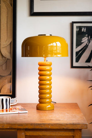 Lifestyle image of the Modern Metal Mustard Table Lamp illuminated and displayed on a wooden desk surrounded by various other home accessories.