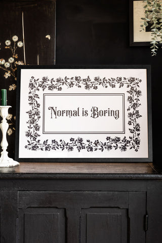 Styled shot of black framed print with 'Normal is Boring' text on a dark background