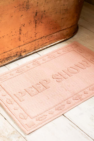 A pink bath mat with the text 'peep show' on, styled on a wooden floor in front of a metal bath.