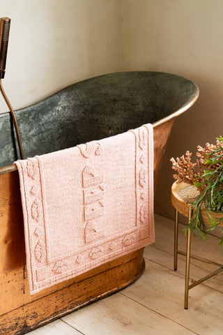 A pink bath mat draped over a metal bath, styled next to a table with a coral ornament and plant on.