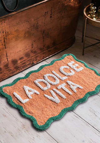 The Peach La Dolce Vita Bath Mat styled on a bathroom floor next to a copper bath and side table with candlesticks on.
