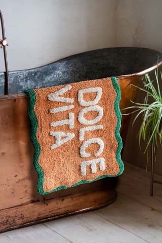 The Peach La Dolce Vita Bath Mat styled draped over the edge of a copper bath, next to a plant.