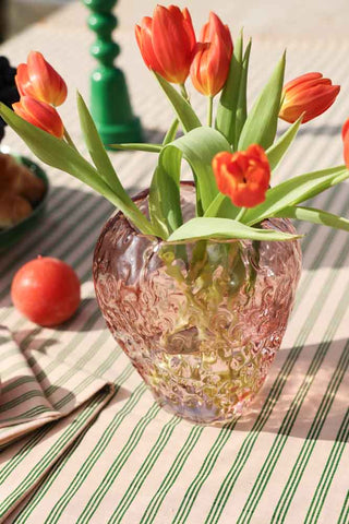 Lifestyle image of the Pink Strawberry Glass Vase displayed on a stripy tablecloth in the sun with flowers inside. 