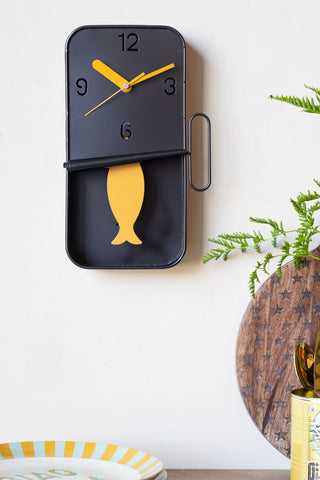 The Quirky Sardine Tin Pendulum Wall Clock on the wall above a table with various dining accessories and a plant on.