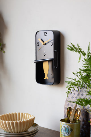 The Quirky Sardine Tin Pendulum Wall Clock on the wall above a wooden table styled with a plant and various dining accessories.