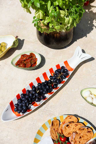 The Red & White Fish-Shaped Ceramic Serving Dish styled with black grapes on, on an outdoor table with other plates/bowls of food and a plant.