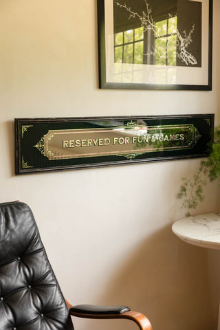 The Reserved For Fun & Games Vintage-Style Mirror displayed on the wall, styled with a chair, table, plant and art print.