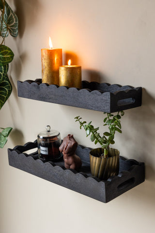 A lifestyle image of The Set Of 2 Black Wooden Scalloped Wall Shelves styled on a neutral wall, with candles and plant accessories