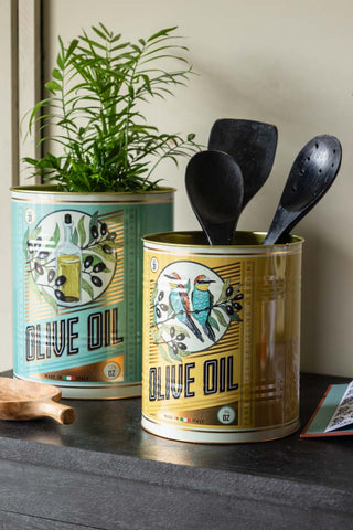 The Set Of 2 Olive Oil Storage Tins - Large displayed together on a black sideboard with utensils and a plant inside.