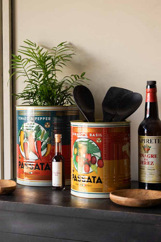 The Set Of 2 Passata Storage Tins - Large styled together on a black sideboard with kitchen accessories and a plant.