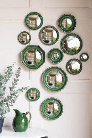The Set of 12 Antique Green & Gold Convex mirror set styled on a light wall, above a table with a green jug and a plant on.