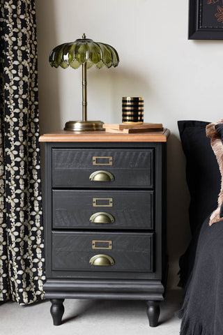 Black bedside table with a lamp and accessories on, styled next to a patterned curtain and bed.