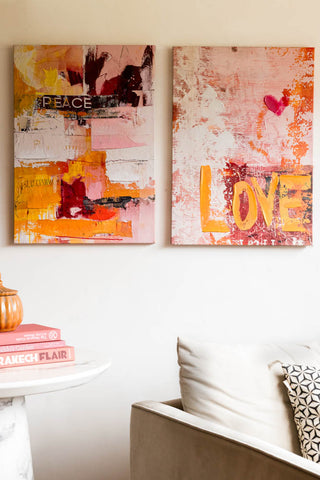 A styled image of the Set of 2 Pink & Orange Abstract Peace/Love Wall Art on a white wall next to an armchair.