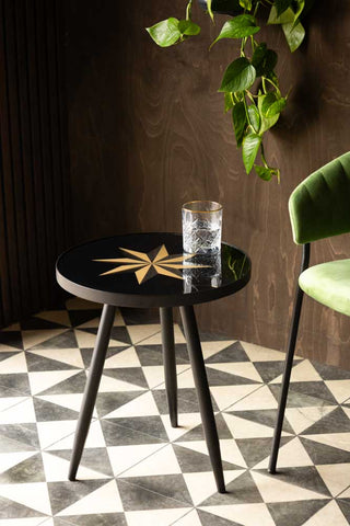 The Small Black Star Side Table with a glass on top, displayed on a geometric floor next to a green chair in front of a dark wooden wall.