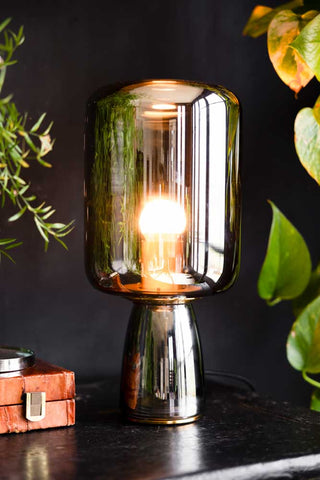 Lifestyle image of the Smoked Table Lamp illuminated and displayed on a black sideboard alongside other home accessories and plants in the background.