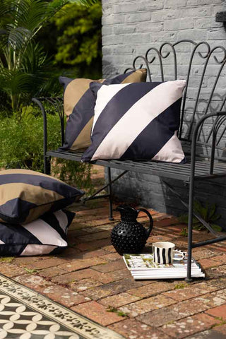 Lifestyle image of the Black & Green Stripe Outdoor Cushion on a garden bench styled with a black jug and a striped mug.