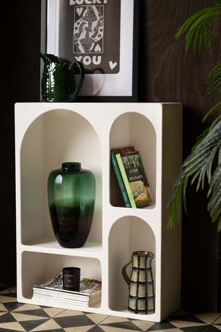 Lifestyle image of the Tall White Alcove Shelf with various accessories displayed inside and on the top, in front of a dark wooden wall with a plant. 