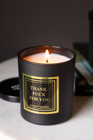 The Thank Fuck For You Scented Candle lit and displayed on a white surface, with the lid and a book in the background.