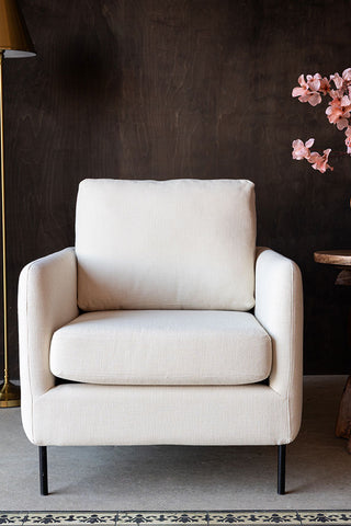 The Edie Natural Armchair seen from the front, with a lamp, side table and flowers in the background.