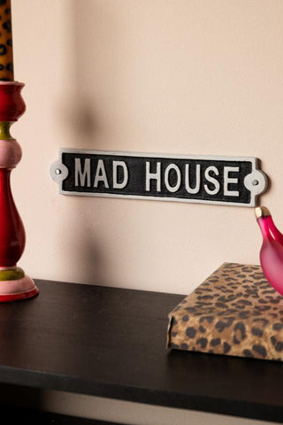 The Madhouse Sign displayed on a neutral wall, above a colourful candlestick holder with a leopard print candle in, a leopard print notebook and a pink banana ornament.